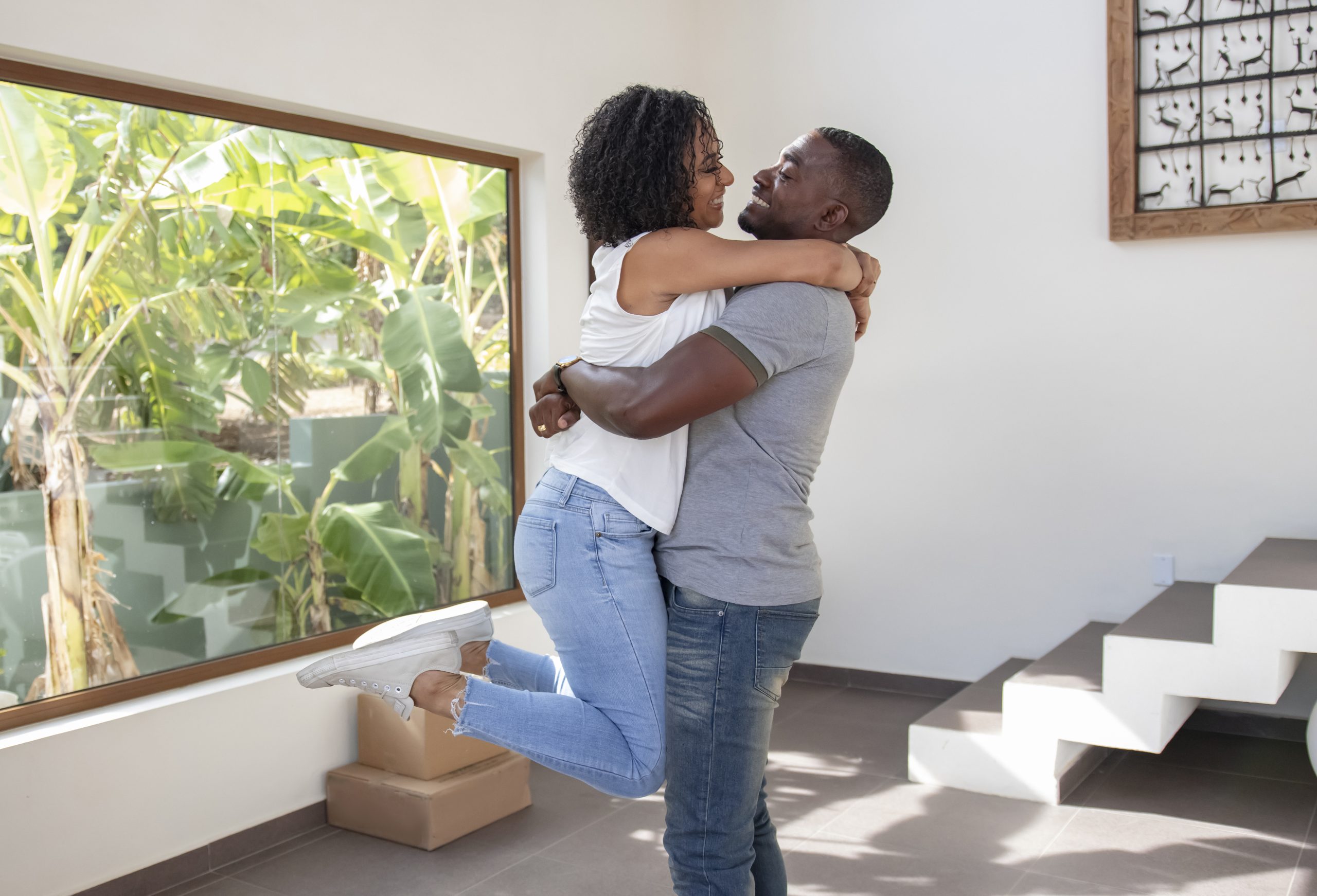 Man and woman are inside a house. The man holds the woman in his arms and they're smiling at each other
